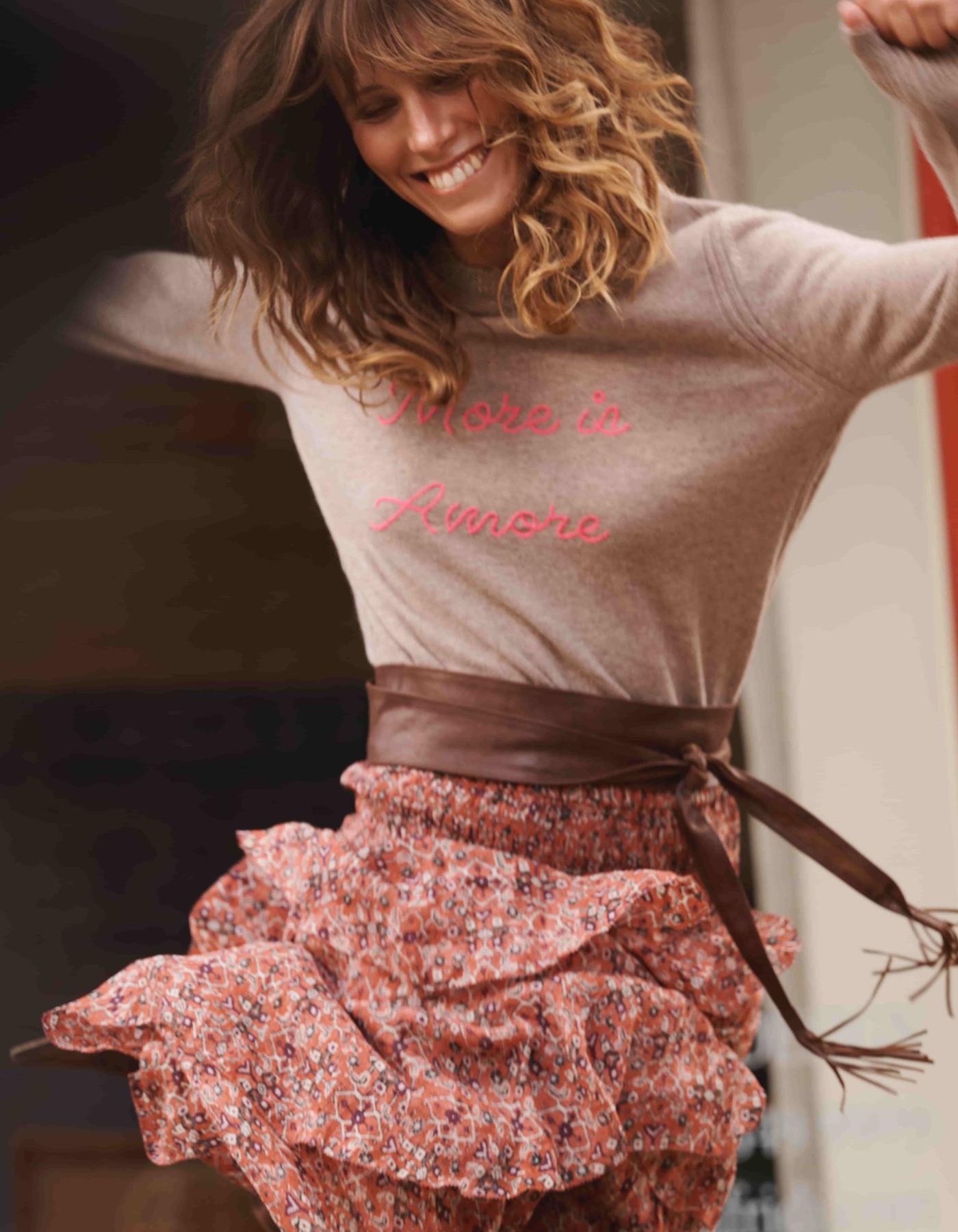 Woman wearing a brown cashmere jumper with text "More is Amore" and a floral skirt,outdoors.