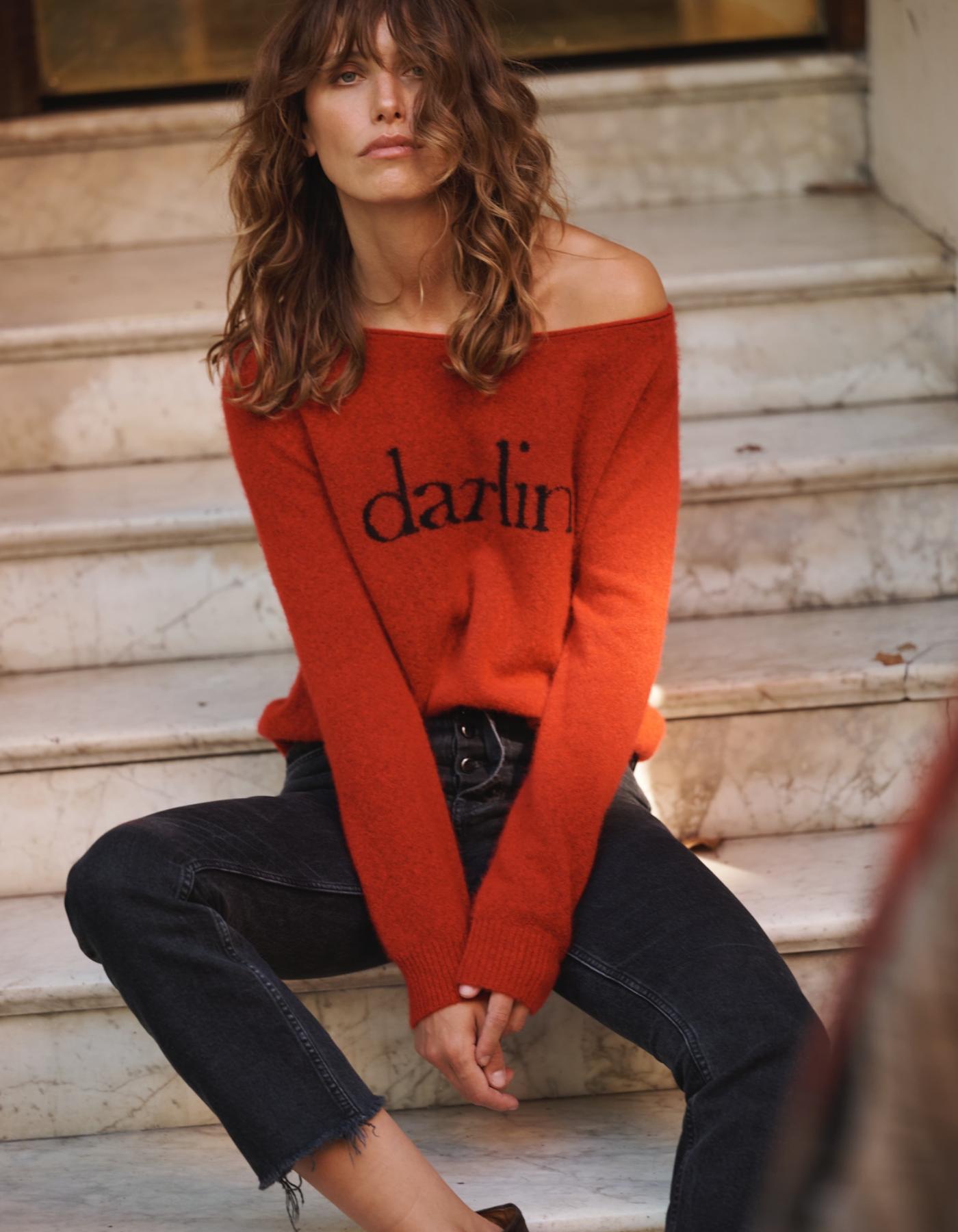 Woman wearing a red sweater with 'darlin' text, sitting on steps.