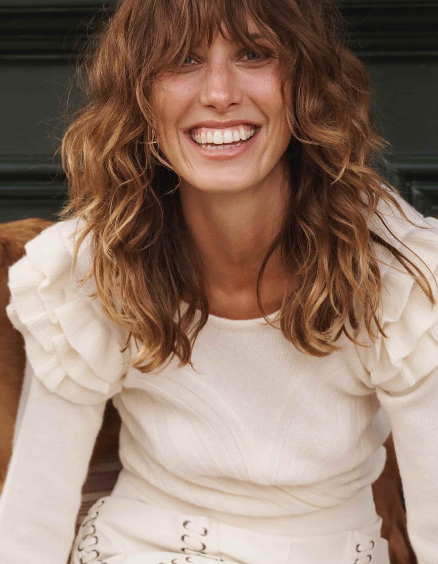 Woman with wavy hair wearing a white cashemere jumper with ruffled shoulders.