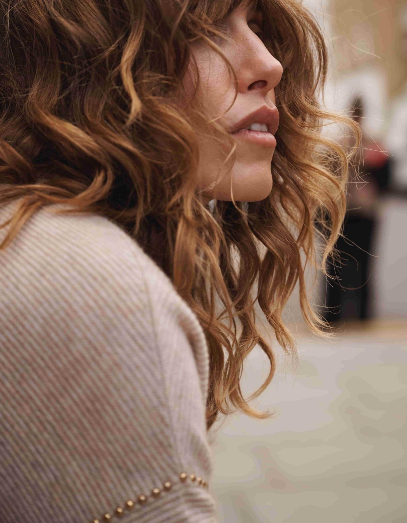 Close-up of a person with wavy hair wearing a beige sweater with gold studs on the sleeve.