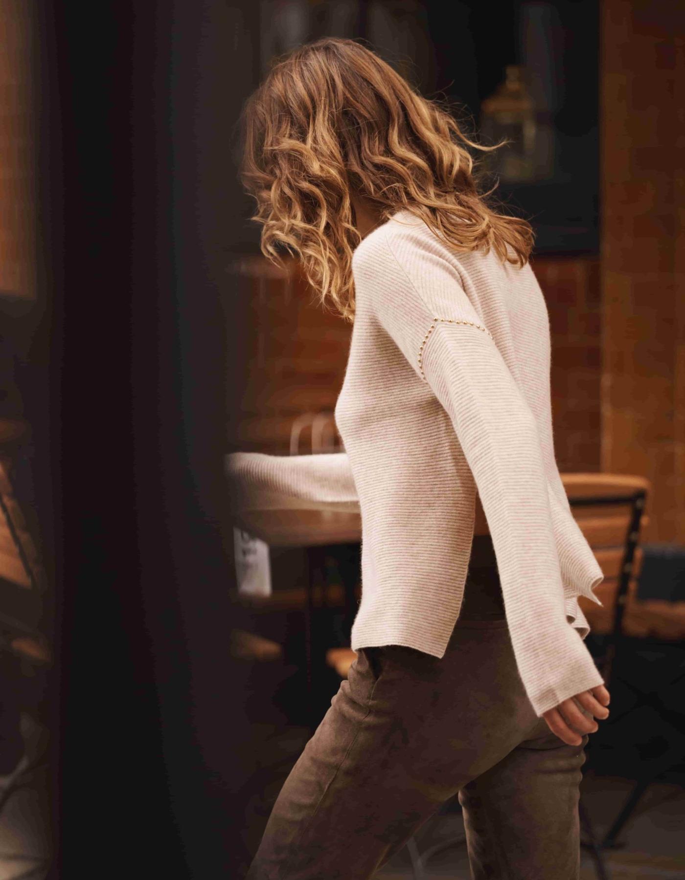 a person with wavy hair wearing a beige cashmere sweater with gold studs on the sleeve.