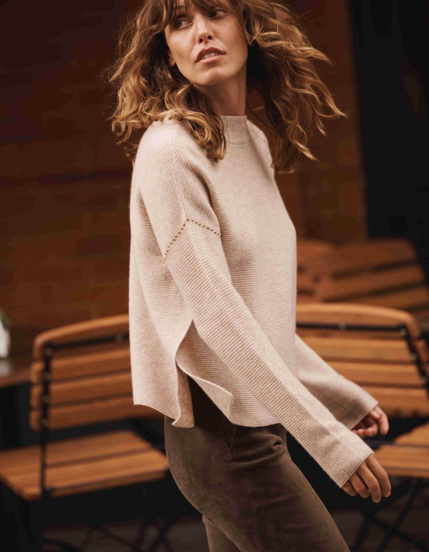Woman wearing a beige sweater in an outdoor setting with wooden chairs.