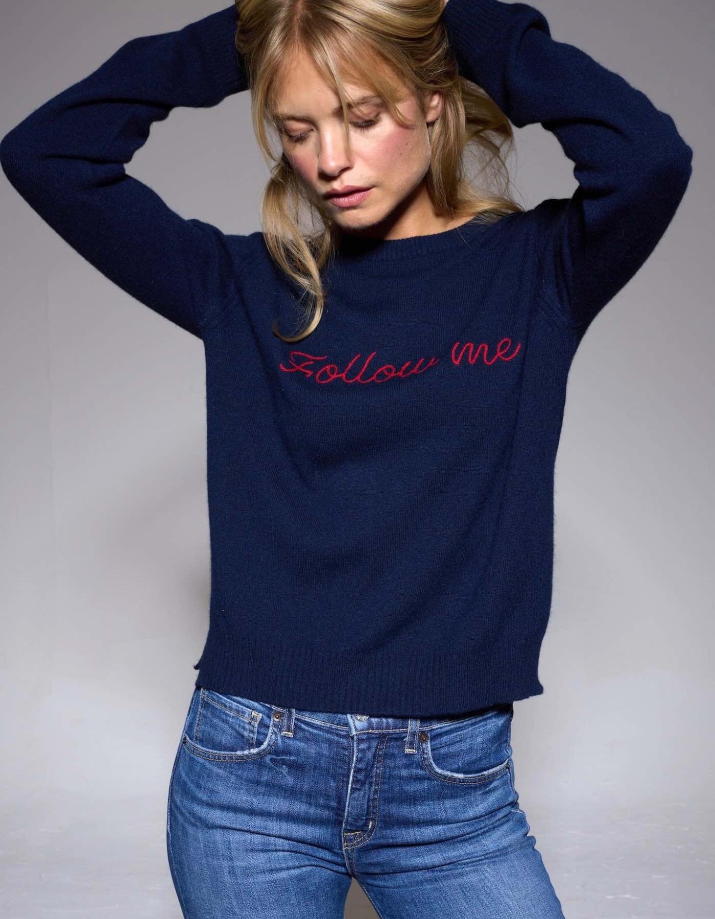 Woman wearing a navy blue sweater with 'Follow me' text and blue jeans on a grey background