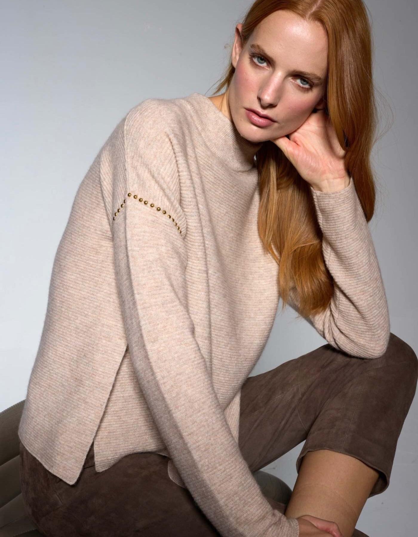 Woman wearing a beige sweater and brown pants against a grey background