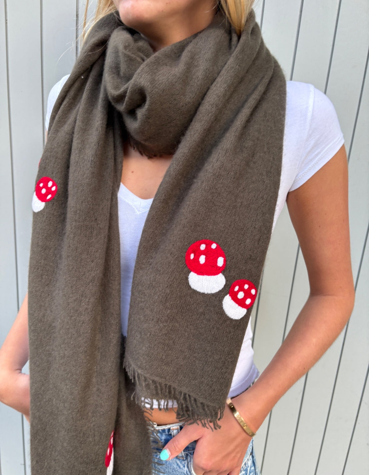 A woman wearing a brown knit scarf with red and white embroidered mushrooms, paired with a white t-shirt and denim jeans, standing against a light gray paneled wall.