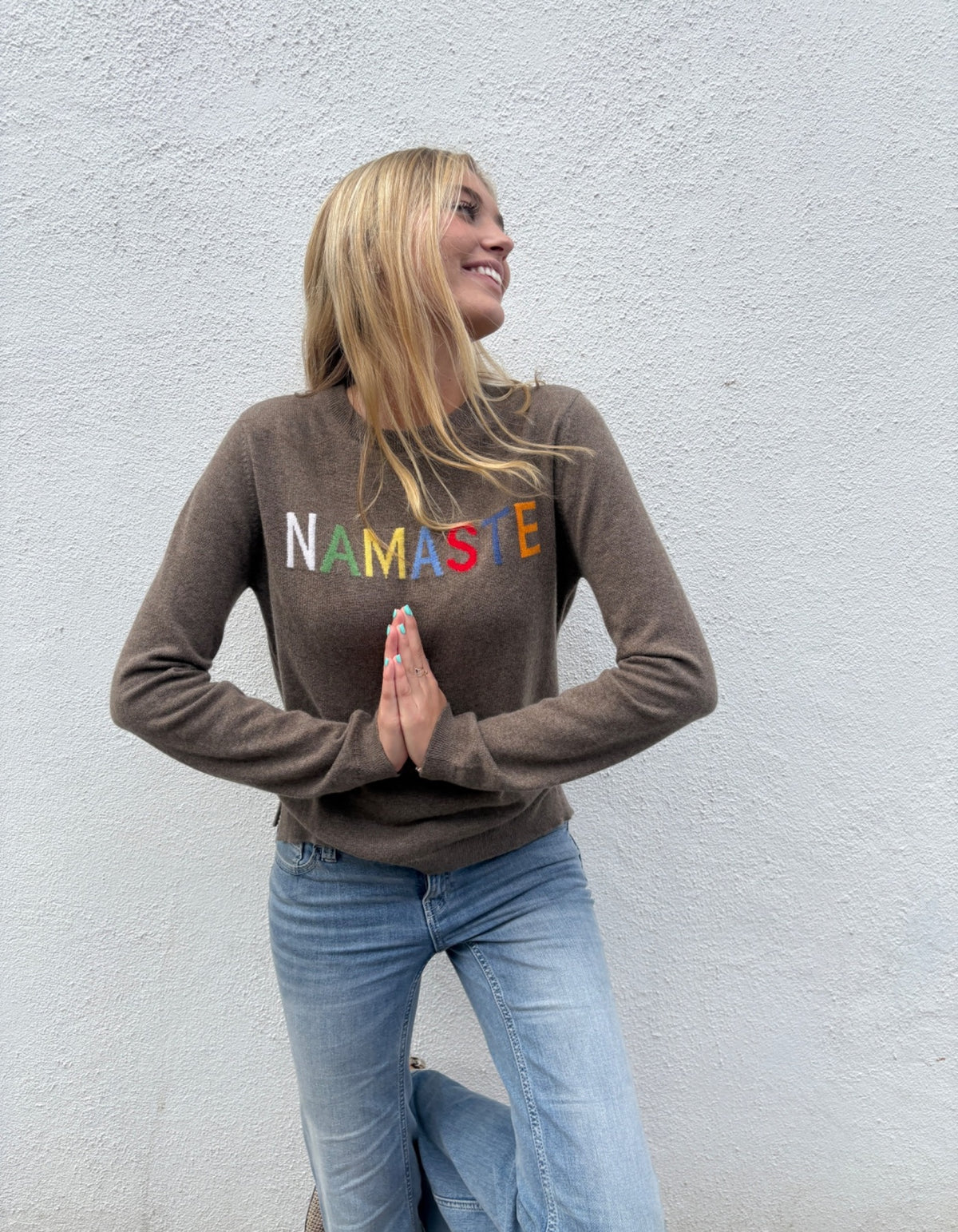 A woman wearing a brown long-sleeve crewneck sweater with 'NAMASTE' printed across the chest in colorful letters, paired with light blue jeans, posing against a textured white wall.