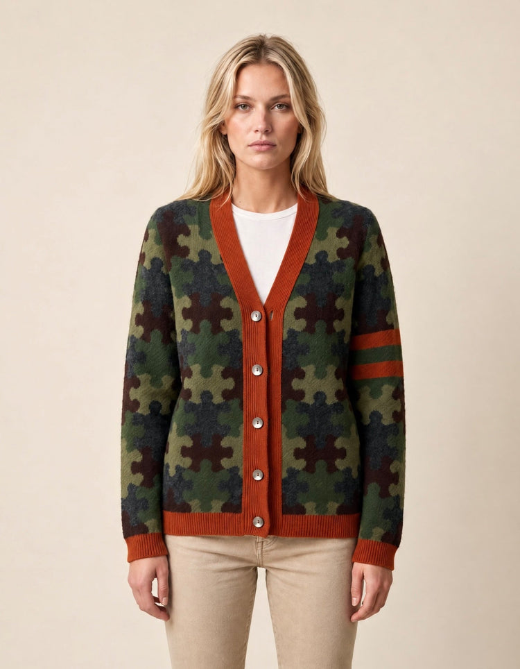 A woman modeling a cardigan sweater with a green and brown puzzle-piece pattern, rust-orange ribbed trim, and a white crewneck shirt underneath, paired with beige pants against a solid beige background.