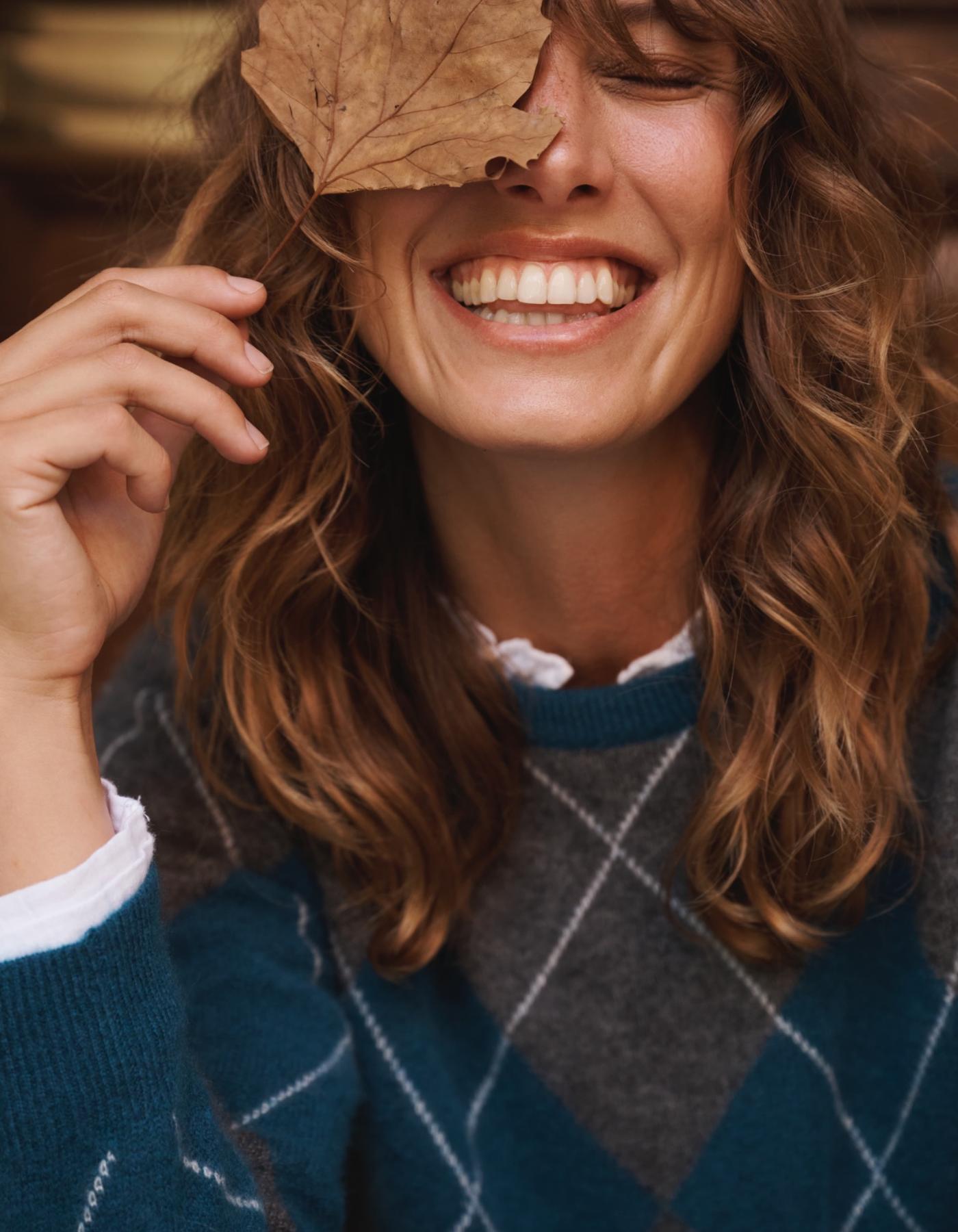 Woman holding a leaf over her eye, wearing a blue and grey cashmere sweater.