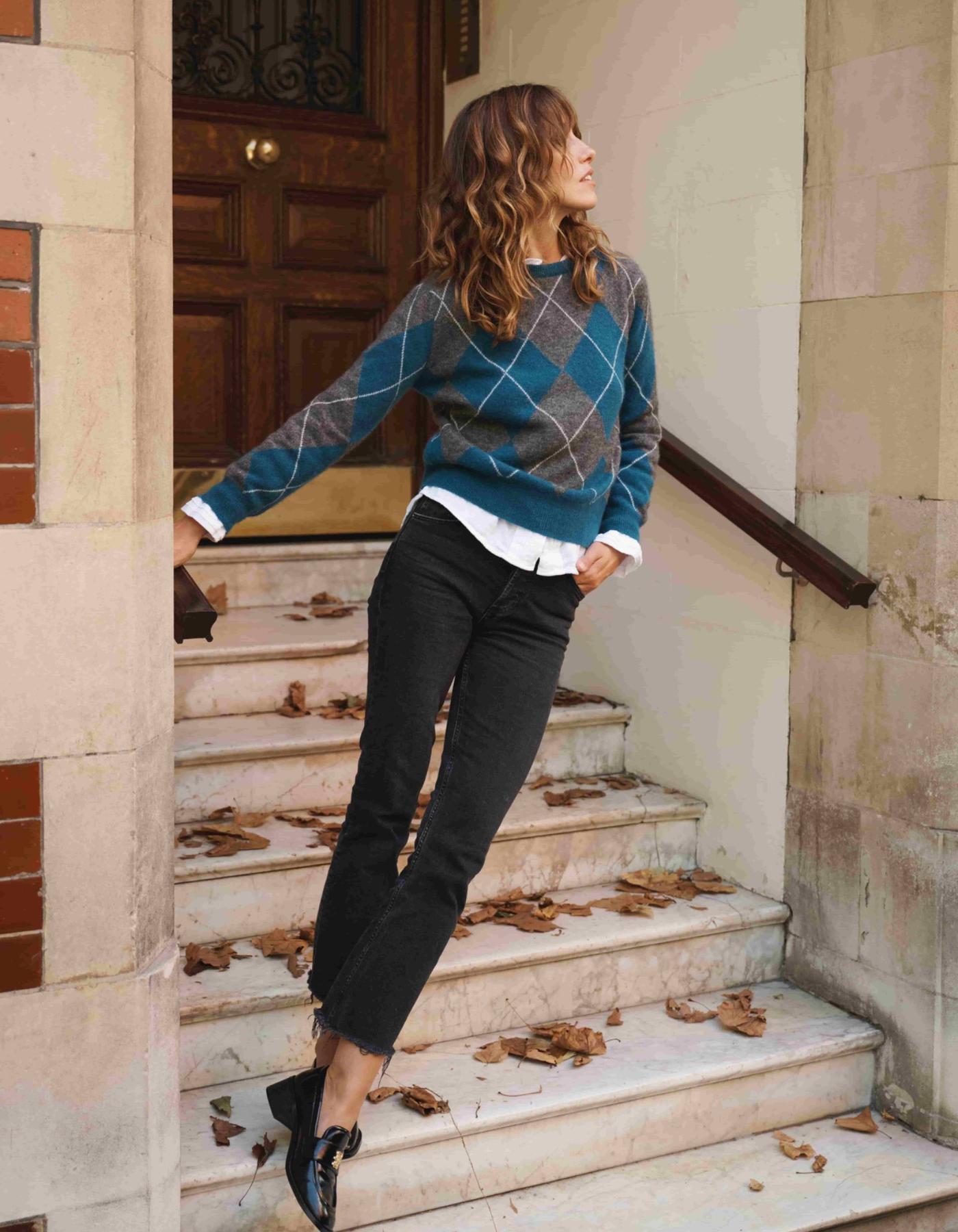 Woman in a blue argyle sweater and black pants standing on stone steps.