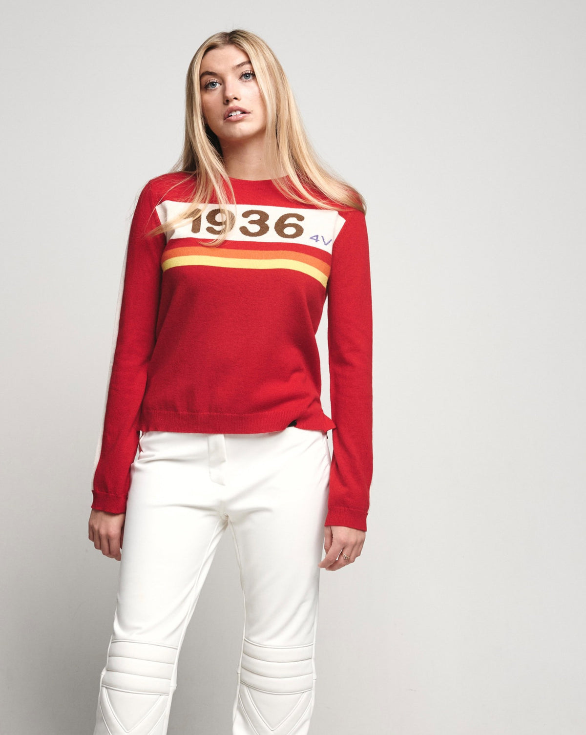 A blonde woman modeling a red long-sleeve crewneck sweatshirt with a white chest graphic reading '1936' and yellow stripes, paired with white pants, against a light gray studio background.