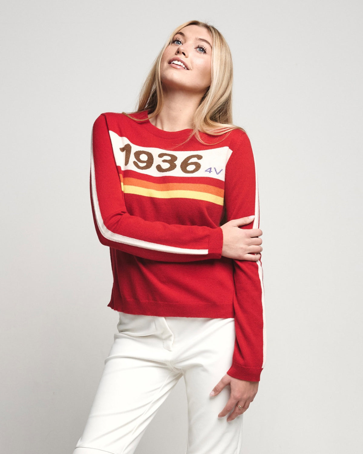 A woman wearing a red long-sleeve knit sweater with a white chest panel, '1936 4v' text, and yellow and orange stripes, paired with white pants, against a light gray studio background.