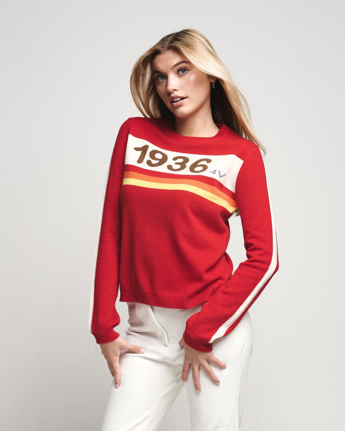 A blonde model wearing a red long-sleeve knit sweater with a white chest graphic reading '1936' and diagonal stripes, paired with white pants, against a light gray studio background.