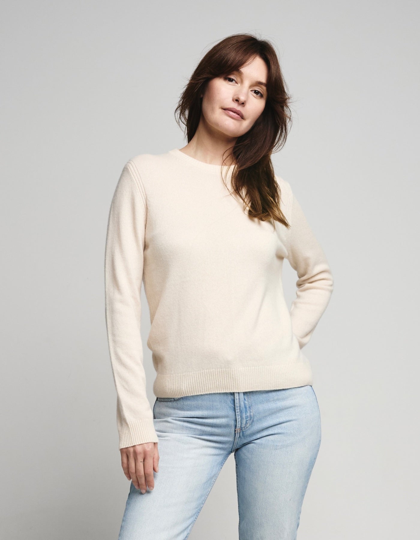 Woman wearing a cream sweater and light blue jeans against a gray background