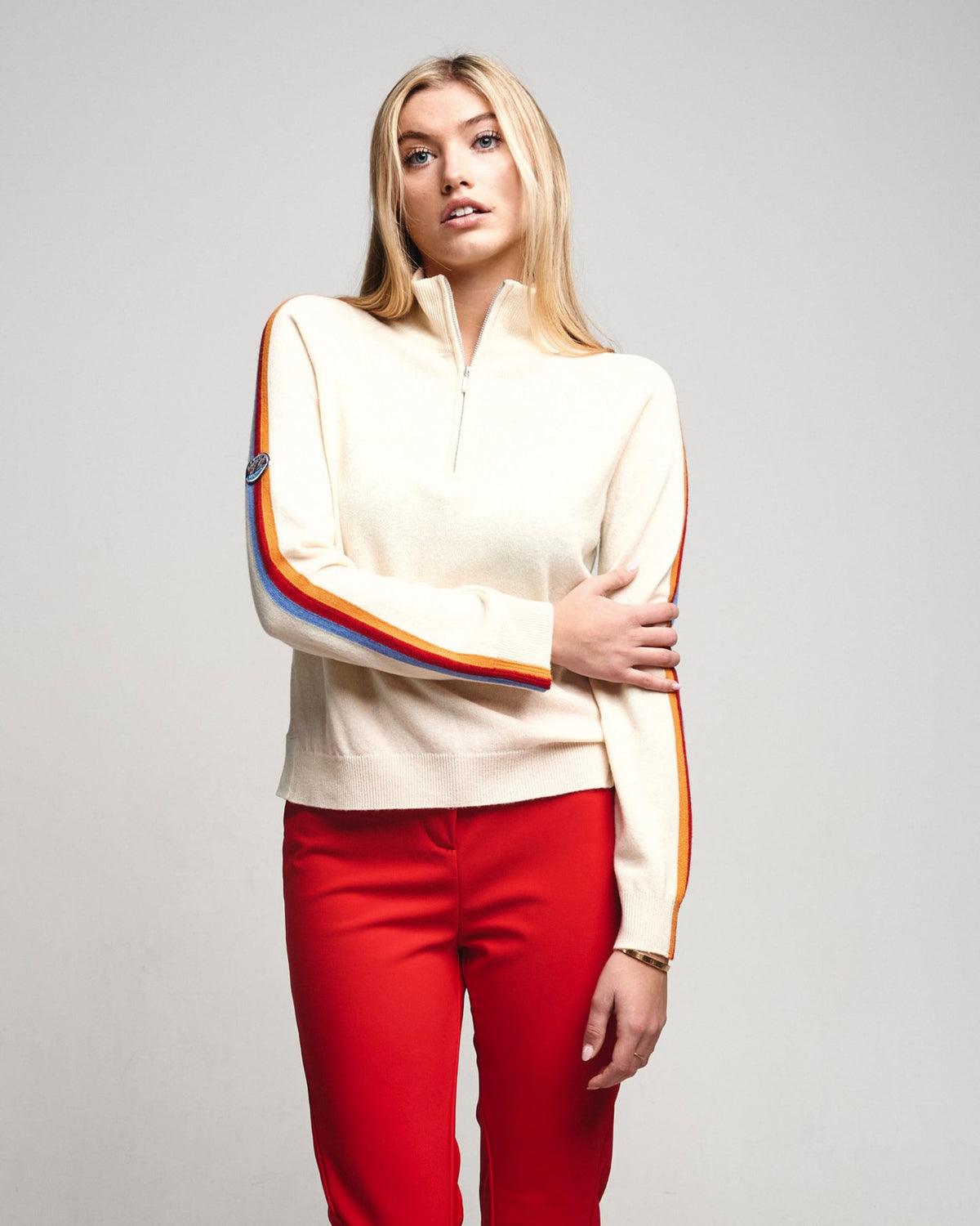 A blonde woman modeling a cream-colored quarter-zip merino chasmere ski jumper with colorful striped ribbed cuffs and a red pair of pants, shot against a solid light gray background.