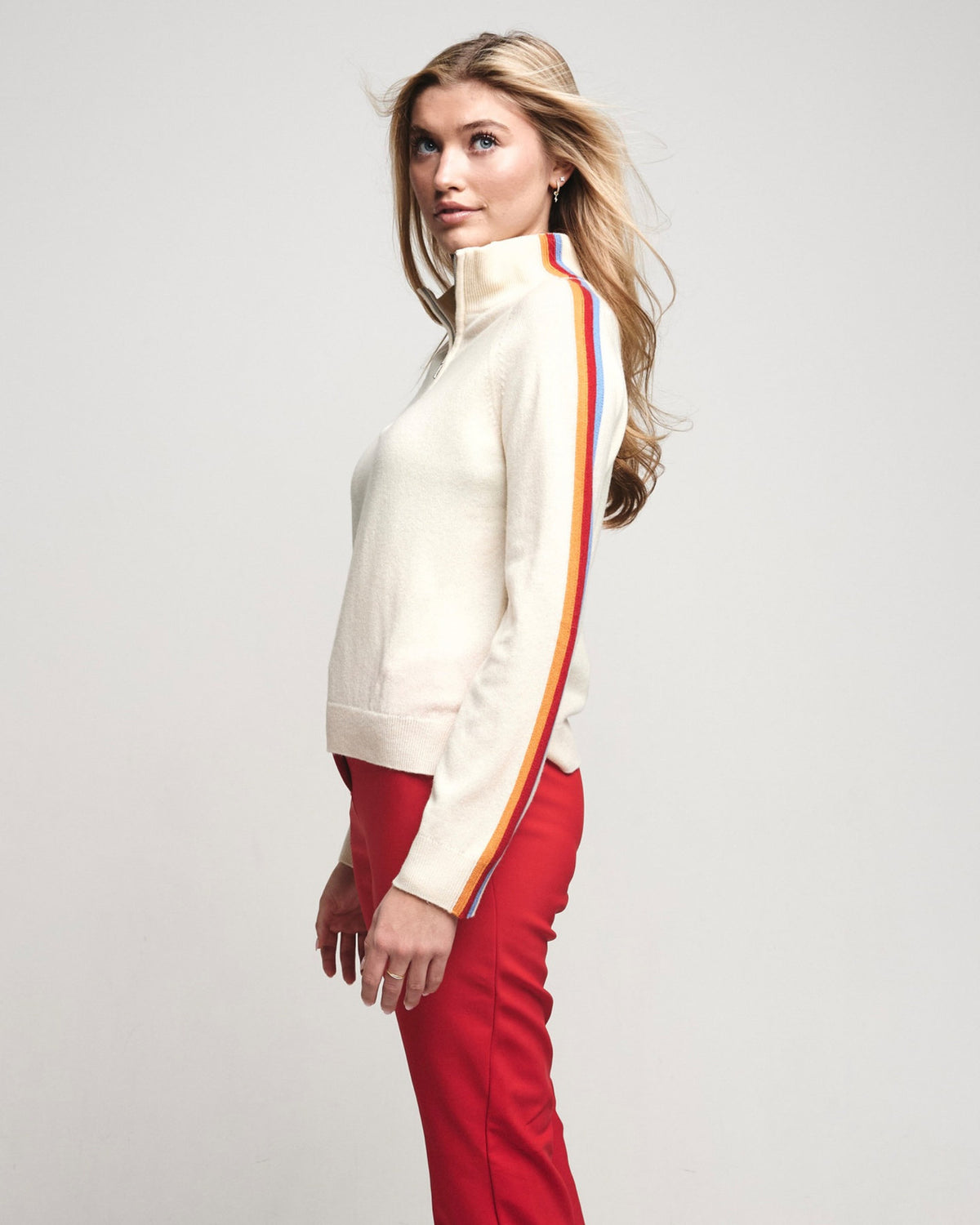 A woman in a cream knit sweater with a red, yellow, and blue stripe down the sleeve, paired with red pants, photographed against a light gray studio background.