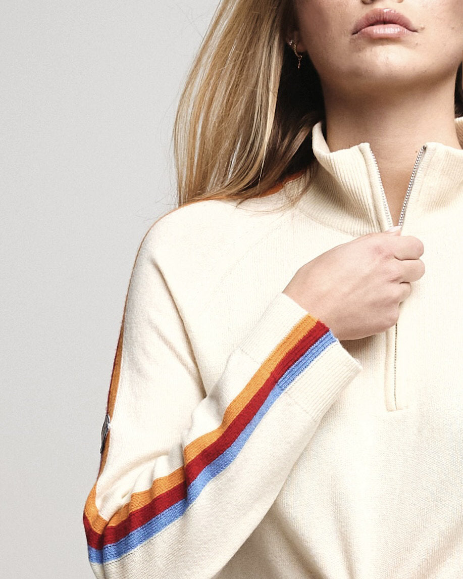 A close-up of a woman wearing a cream-colored quarter-zip sweater with colorful striped ribbed cuffs and sleeves, featuring orange, yellow, red, and blue stripes.