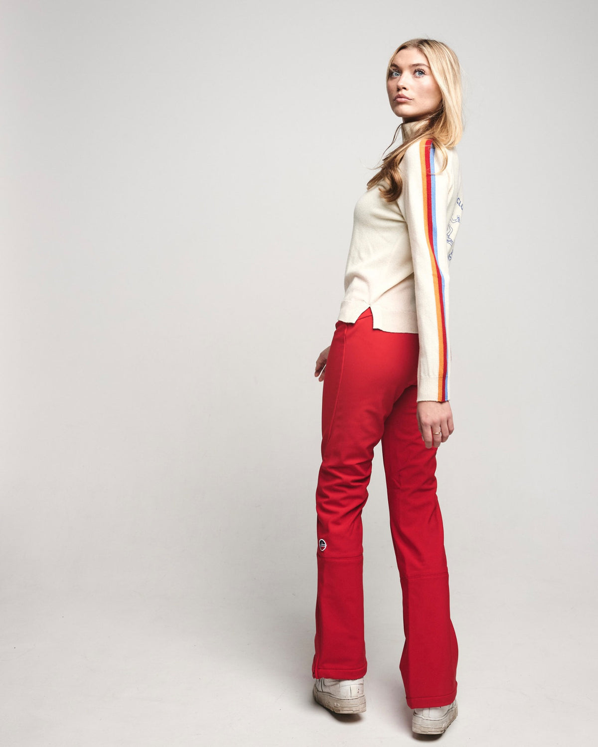 A woman modeling a cream knit sweater with a red, yellow, and orange vertical stripe down the sleeve, paired with bright red flared pants and metallic silver shoes, photographed against a light gray studio background.