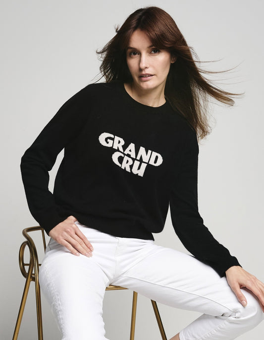 Woman wearing a black cashmere sweater with 'GRAND CRU' text, sitting on a chair against a plain background
