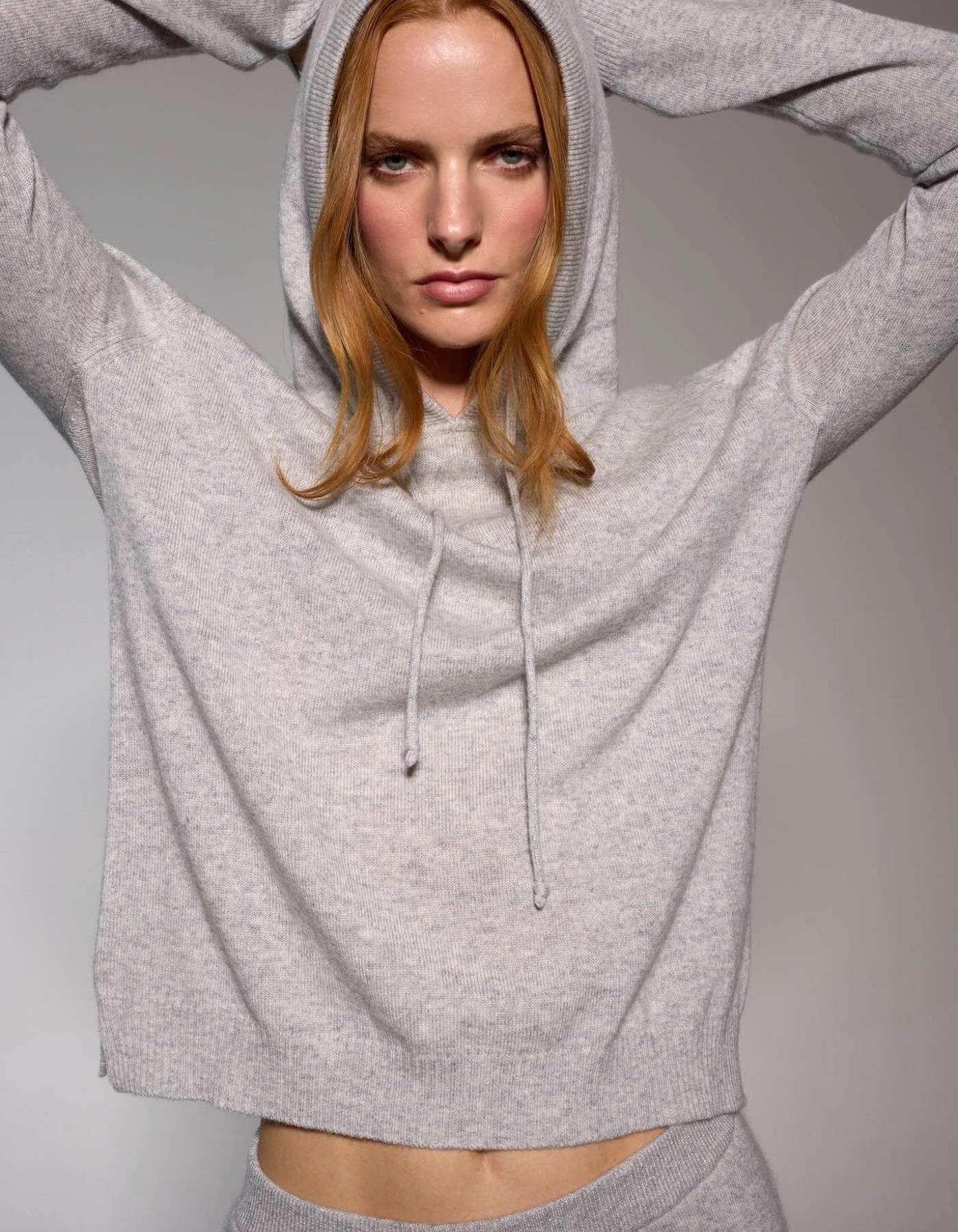Woman wearing a grey chasmere hoodie with arms raised against a plain background