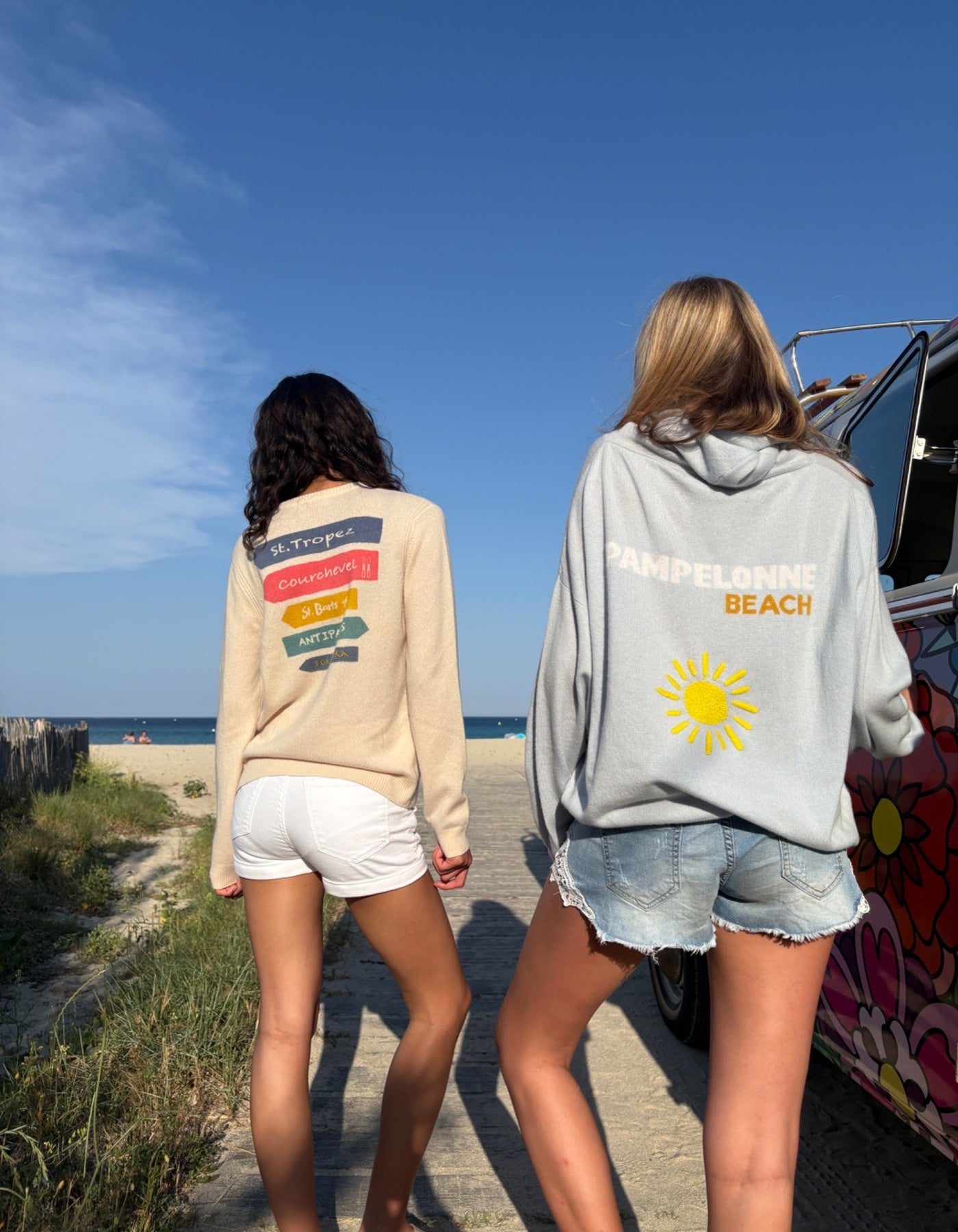 Two people walking on a boardwalk by the beach, wearing cashmere jumper and hoodies with visible text and graphics.
