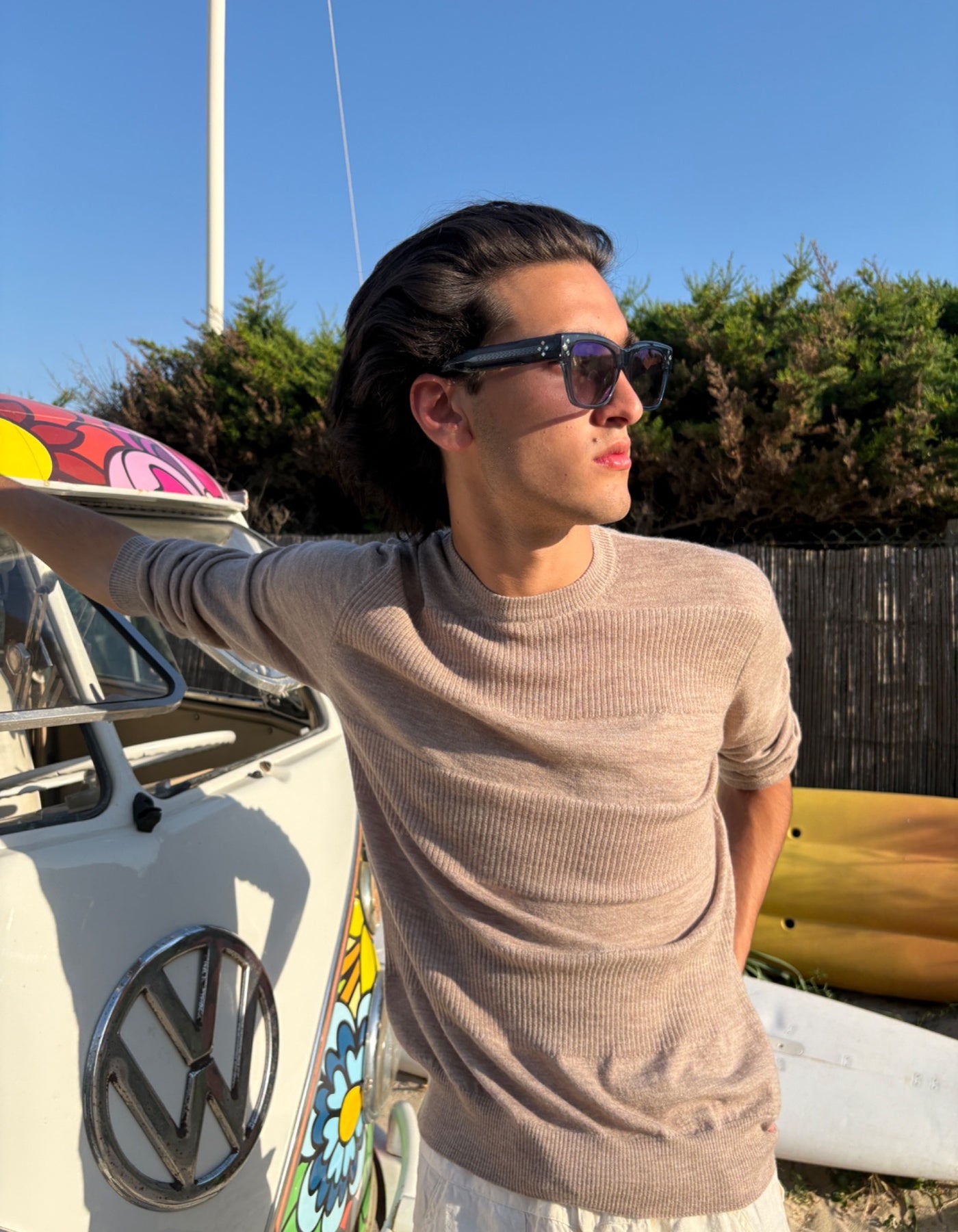 Man wearing sunglasses and a beige cashmere sweater standing next to a colorful surfboard on the beach