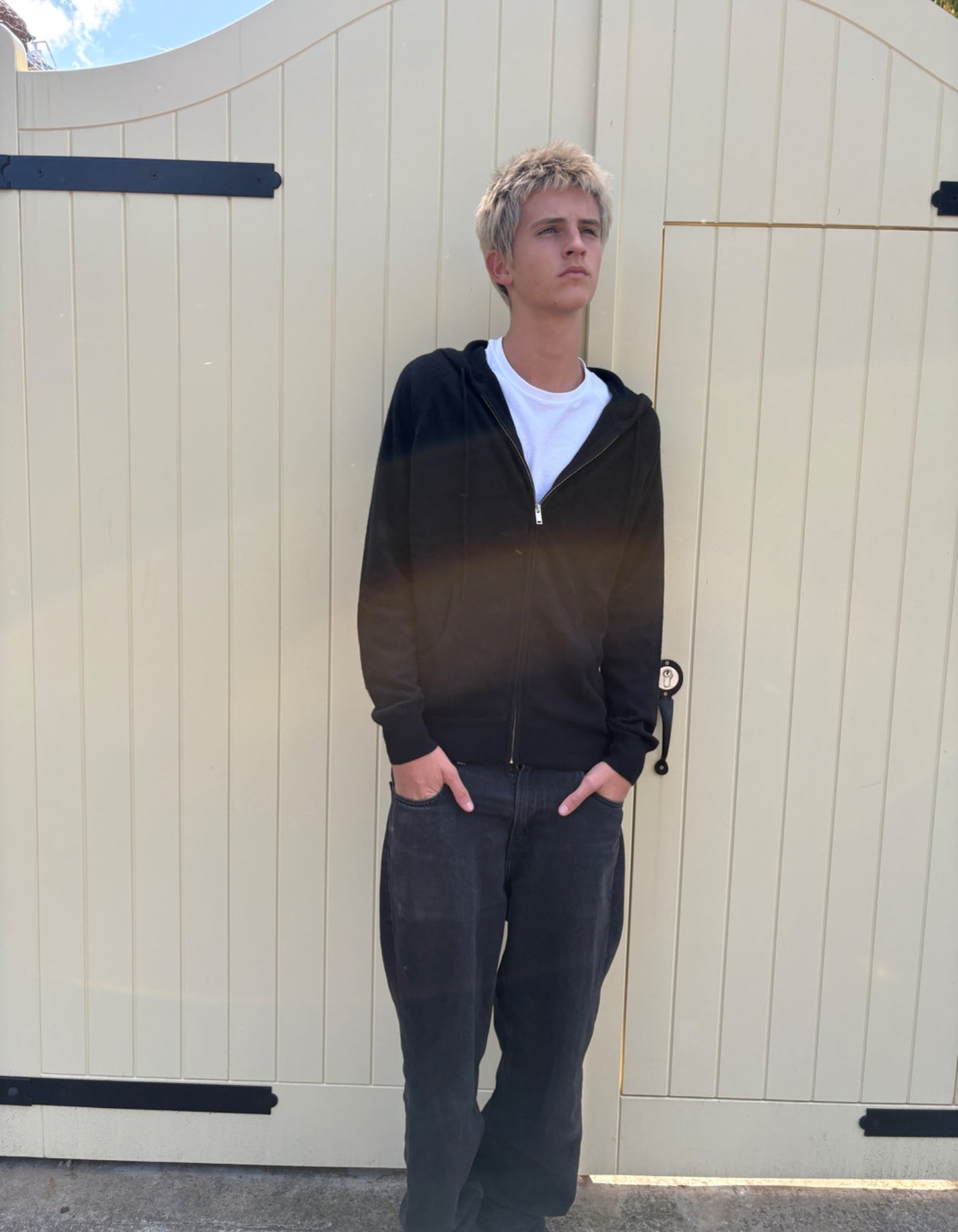 Person wearing a black hoodie and dark pants standing in front of a beige gate.