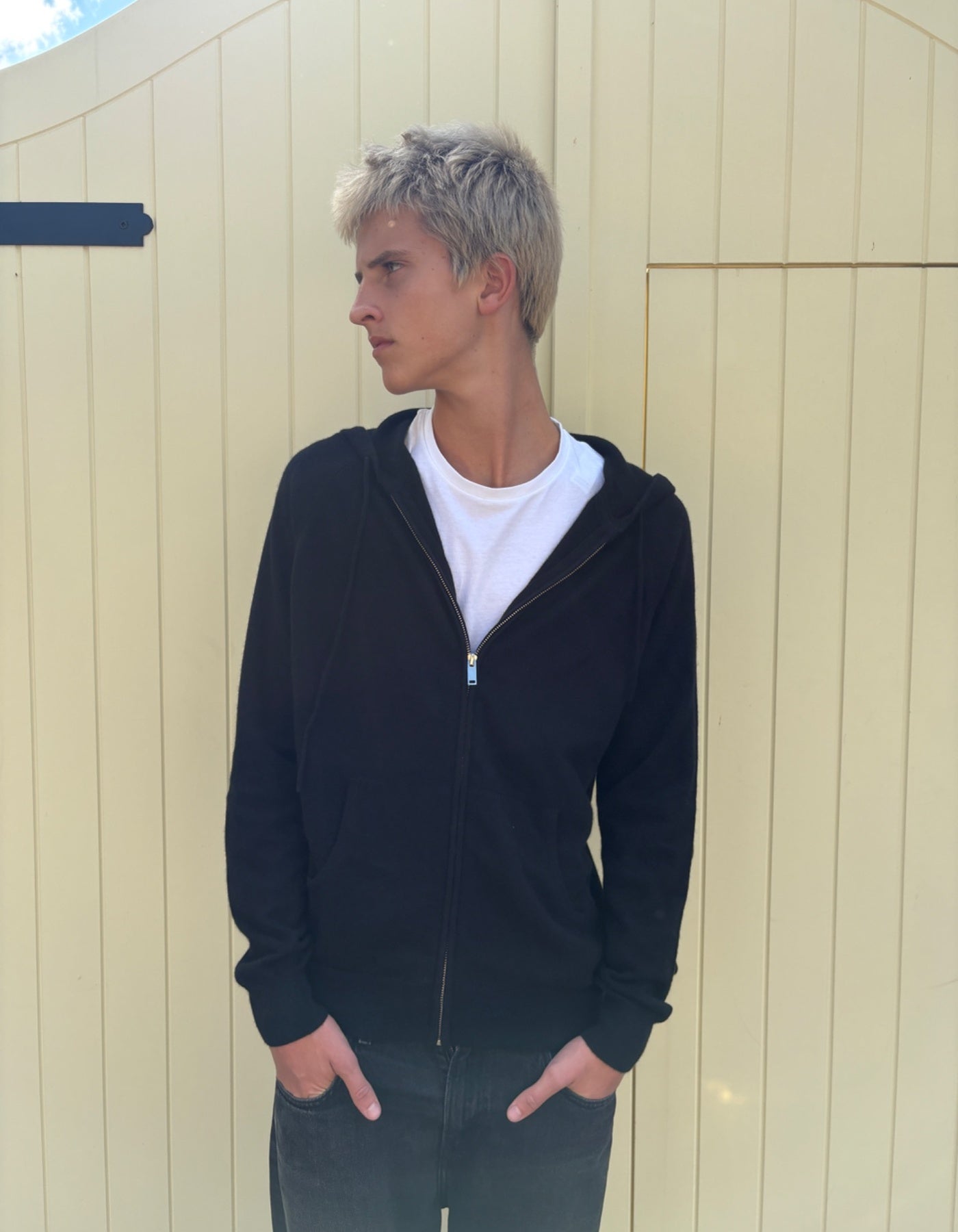 Person wearing a black hoodie and white shirt standing against a beige wall.