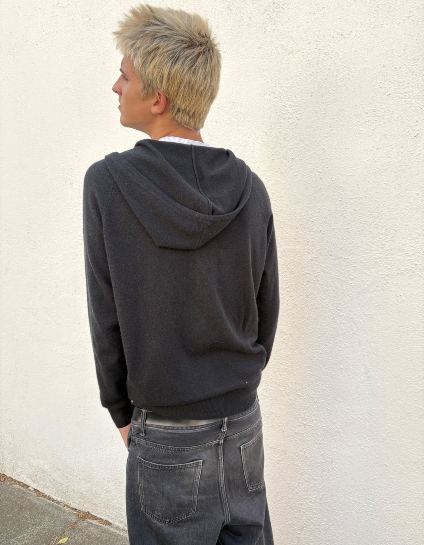 Person wearing a dark hoodie and jeans against a light wall.