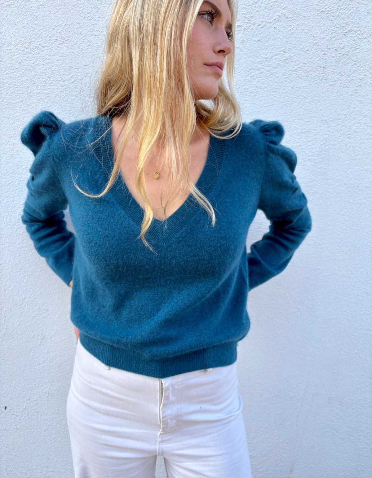 A woman wearing a teal blue knit sweater with puffed shoulders and a V-neck, paired with white pants, standing against a light blue textured wall.