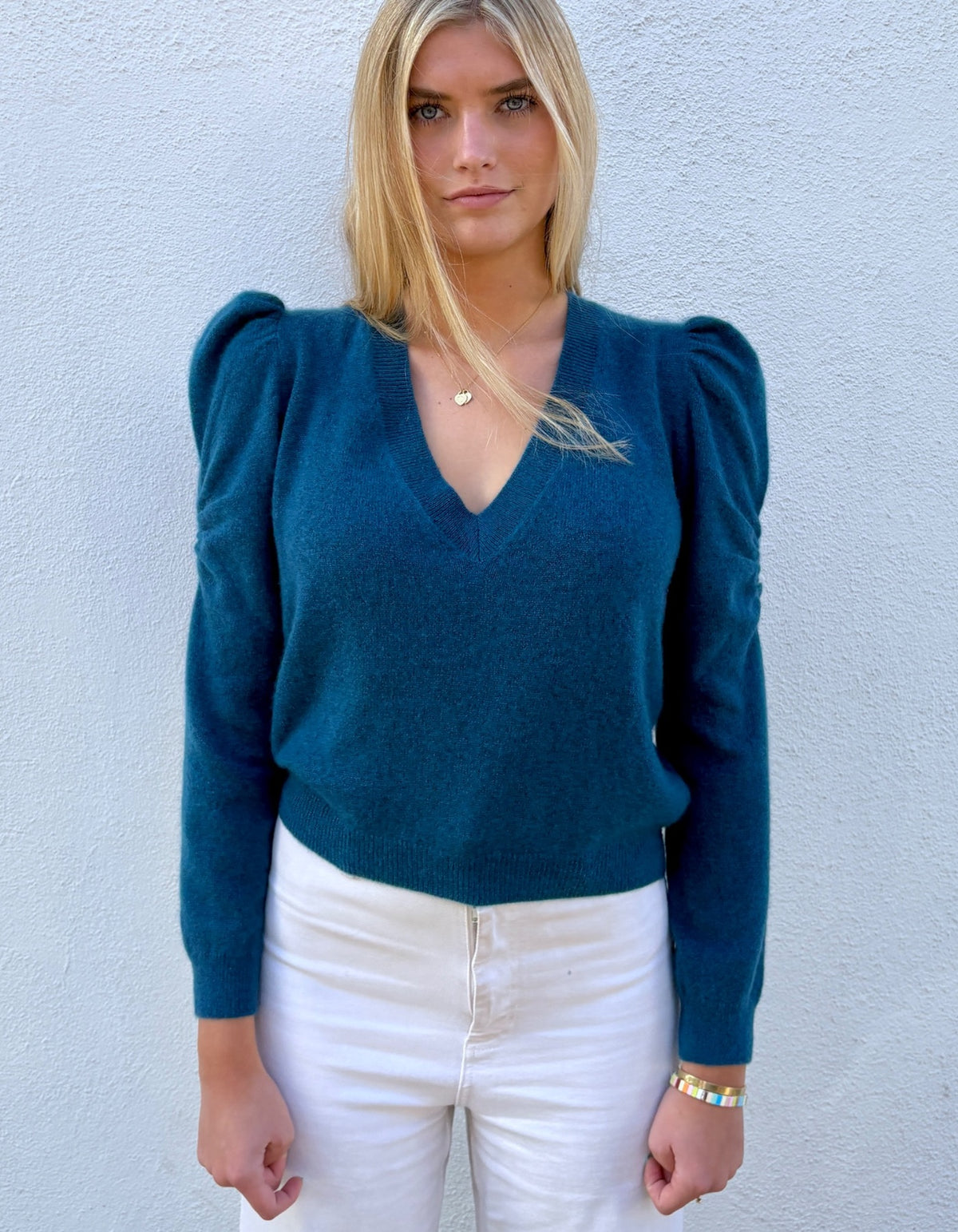 A woman wearing a teal blue long-sleeve knit sweater with puffed shoulders and a V-neck, paired with white pants, standing against a light blue textured wall.