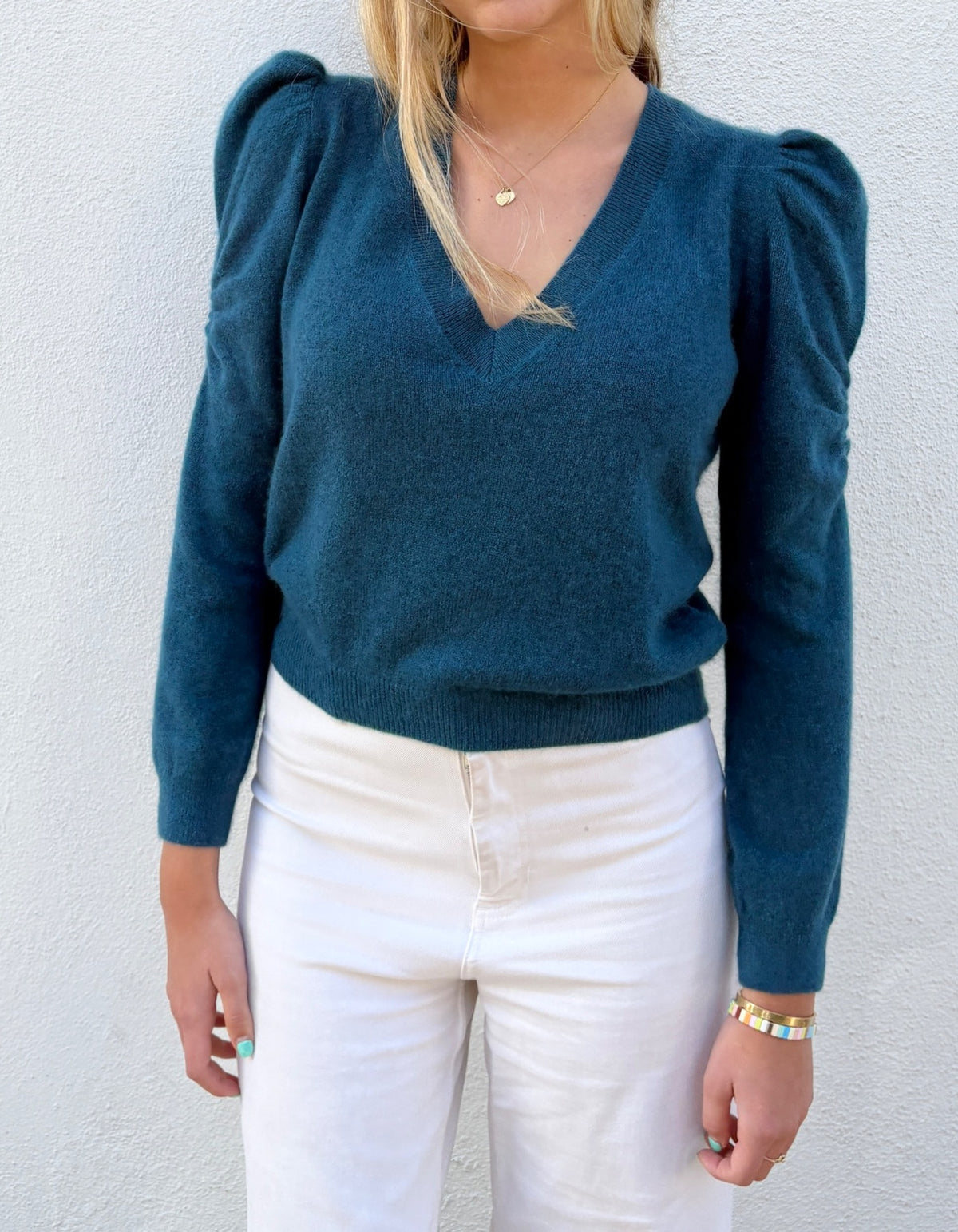 A woman wearing a teal blue long-sleeve knit sweater with puffed shoulders and white pants, standing against a light textured wall.