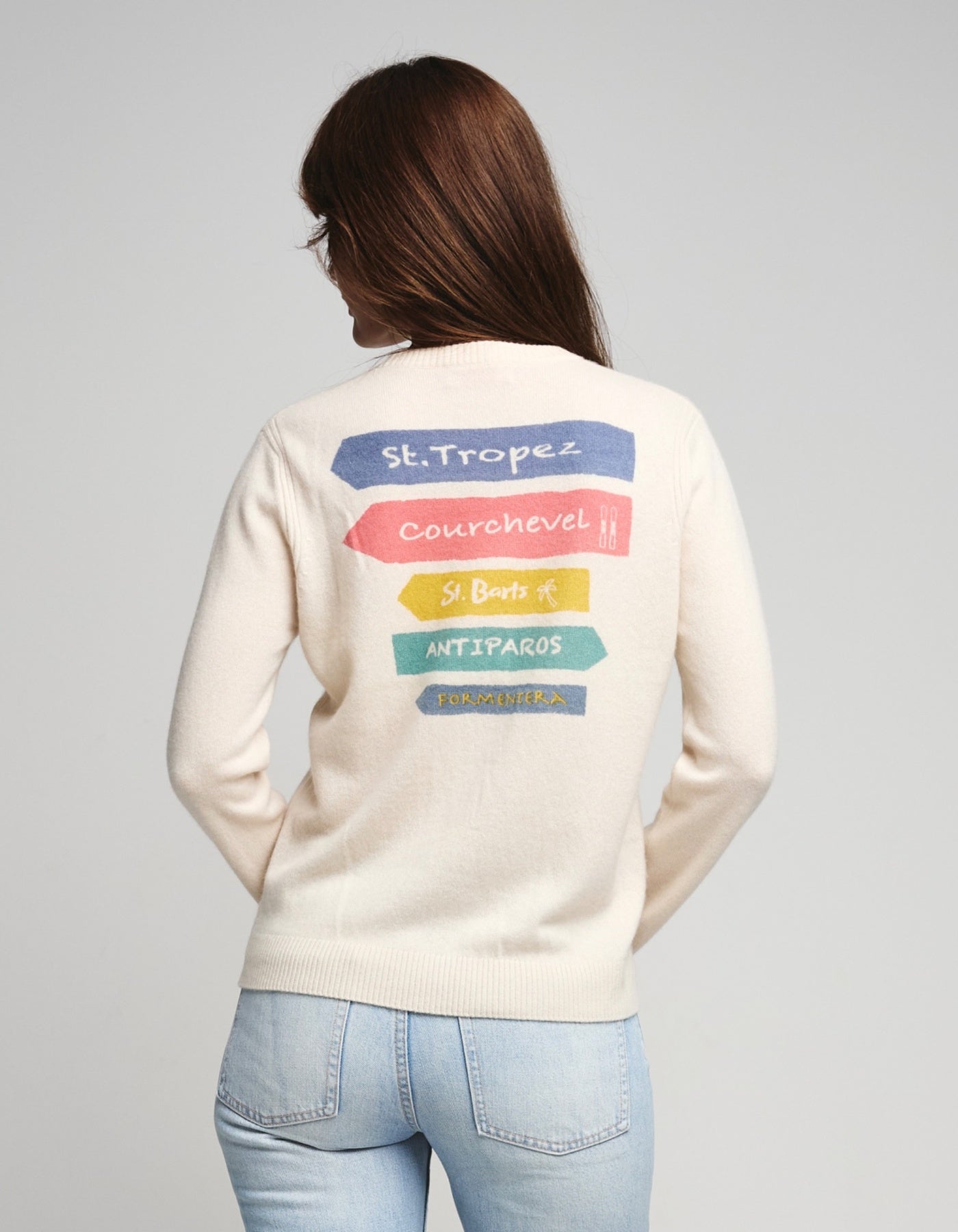 Person wearing a cream-colored sweater with colorful text on the back saying St. Tropez, Courchevel, St. Barts, Antiparos, Formentera against a white  background