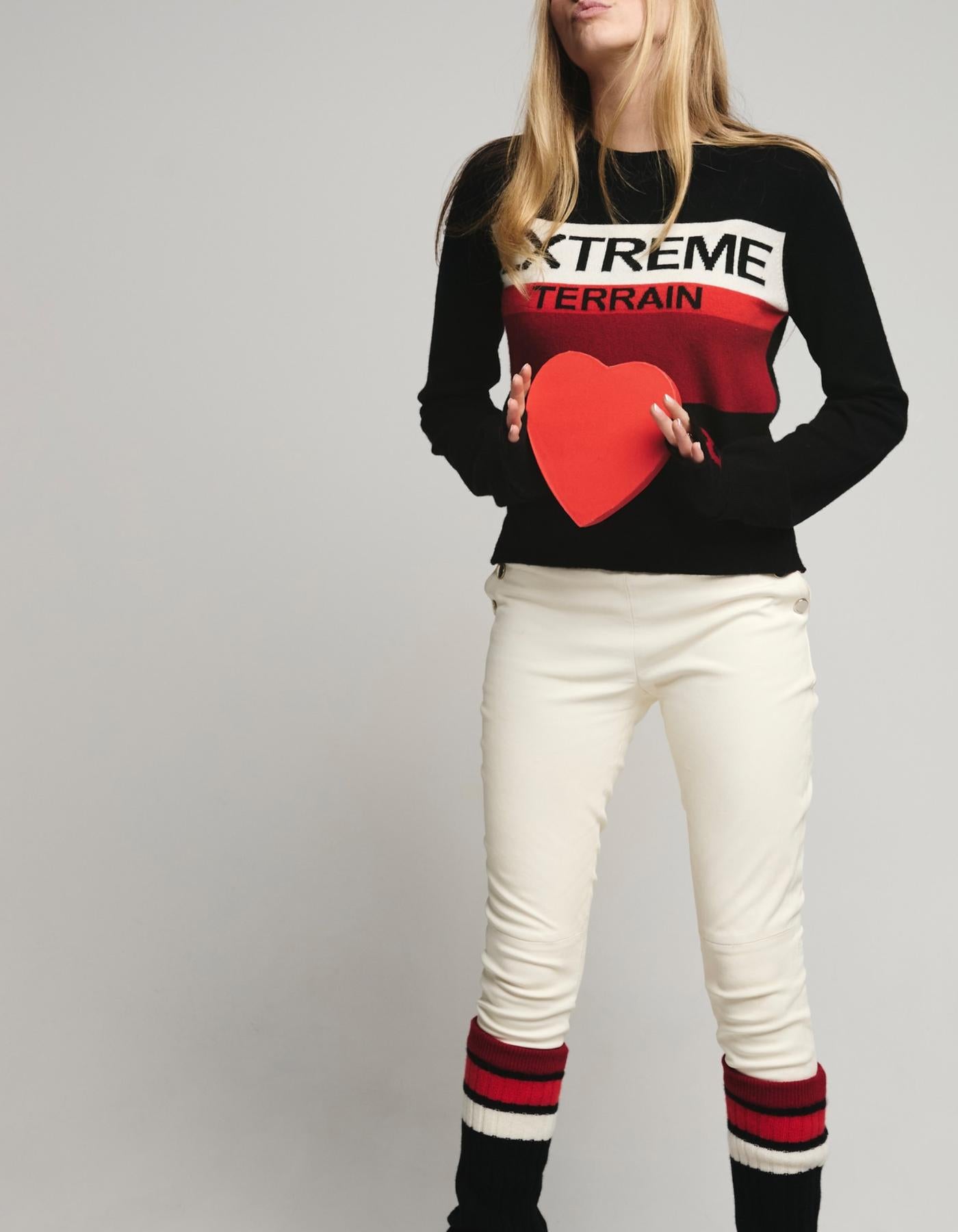 Person wearing a black sweater with 'Extreme Terrain' text and a red heart, white pants, and black socks with red stripes on a gray background.