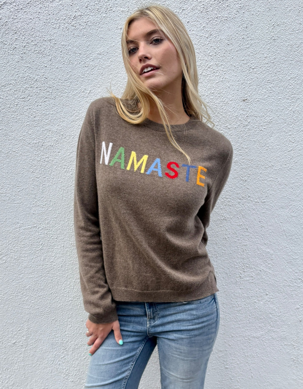 A blonde woman wearing a brown crewneck sweatshirt with the word 'NAMASTE' printed across the chest in colorful block letters, paired with blue jeans, standing against a textured light gray wall.