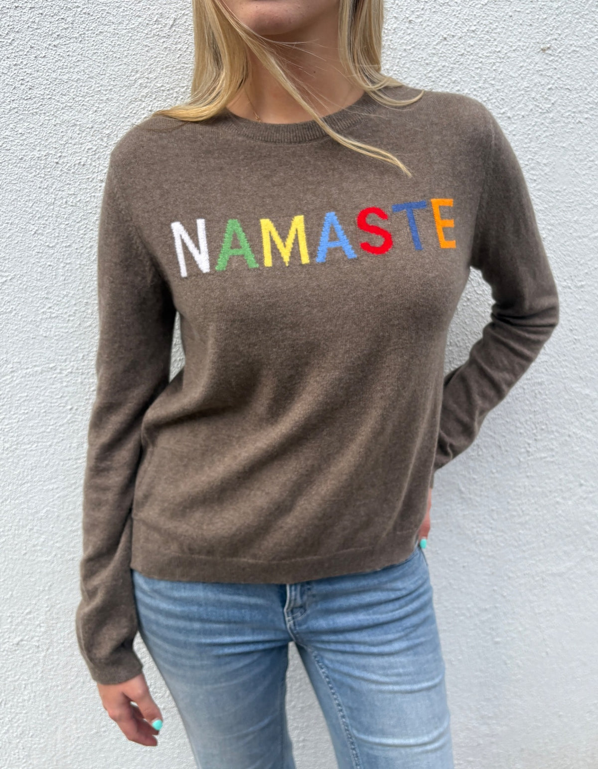 A woman wearing a brown knit sweater with the word 'NAMASTE' spelled out in colorful letters across the chest, paired with light blue jeans, standing against a textured white wall.