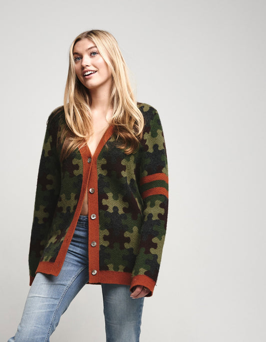 A woman modeling a green and black puzzle-piece camouflage cardigan with rust-orange trim and button-front closure, paired with light blue jeans against a light gray studio background.