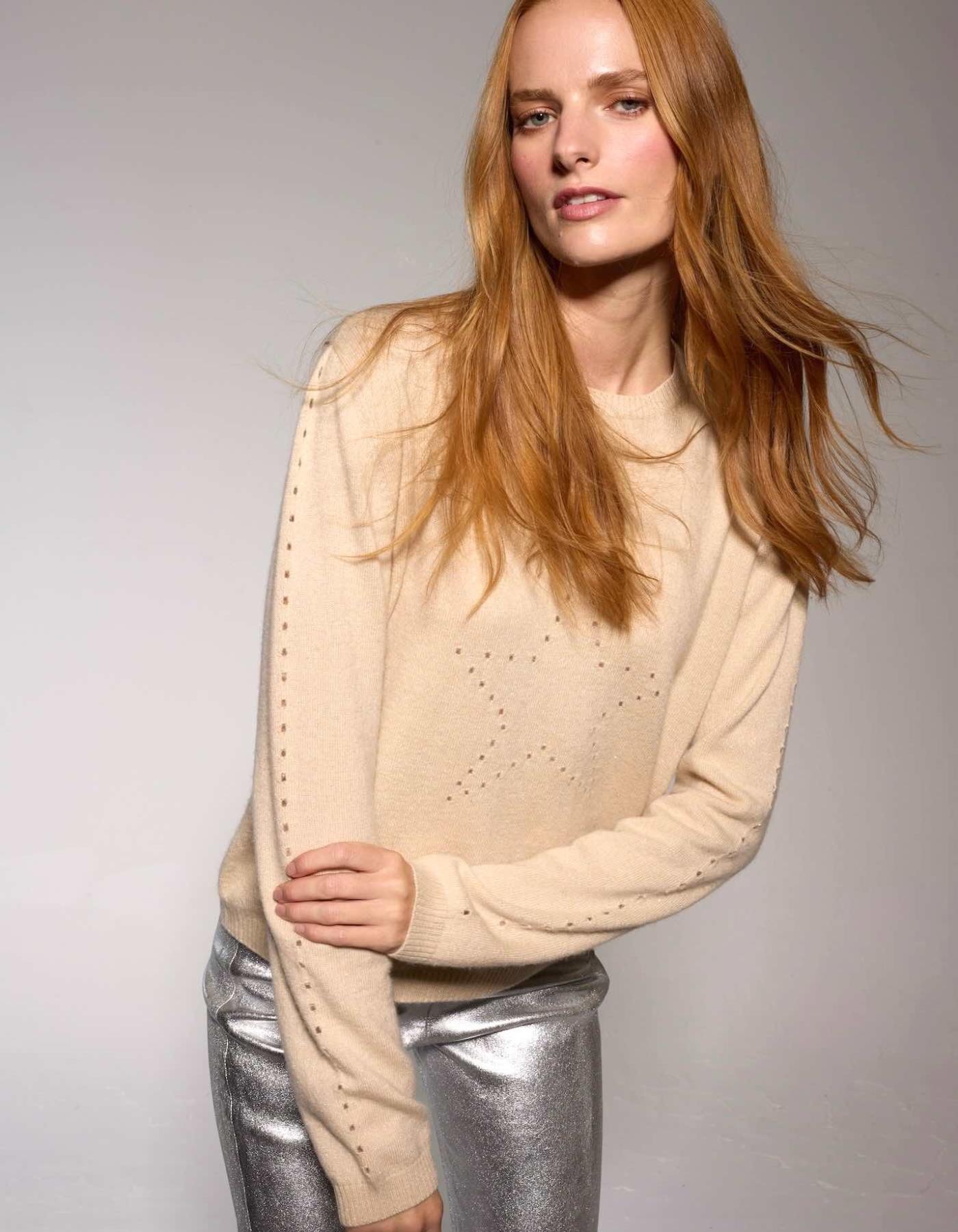 Woman wearing a beige sweater with dropstitich shaping a star and silver pants against a plain background