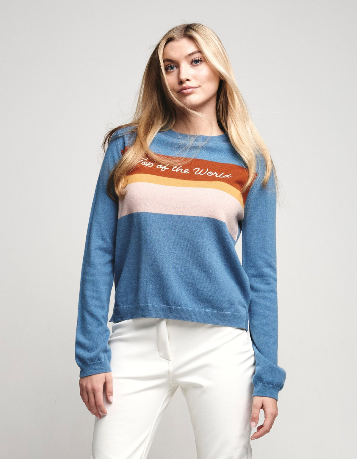 A woman modeling a blue crewneck sweater with horizontal stripes in orange, beige, and white across the chest, featuring the text 'Too of the World' in white script. She is wearing white pants against a plain light gray studio background.