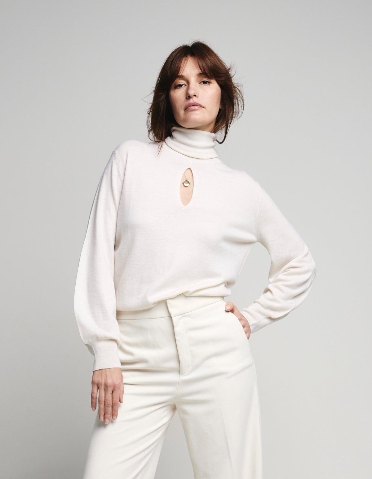 A model wearing a cream-colored turtleneck sweater with a small central cutout and a beige oval patch, paired with matching cream trousers, against a light gray studio background.