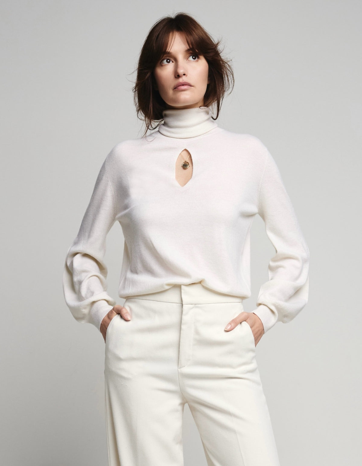 A model wearing a cream-colored knit turtleneck sweater with a keyhole cutout and matching cream trousers, standing against a light gray studio background.