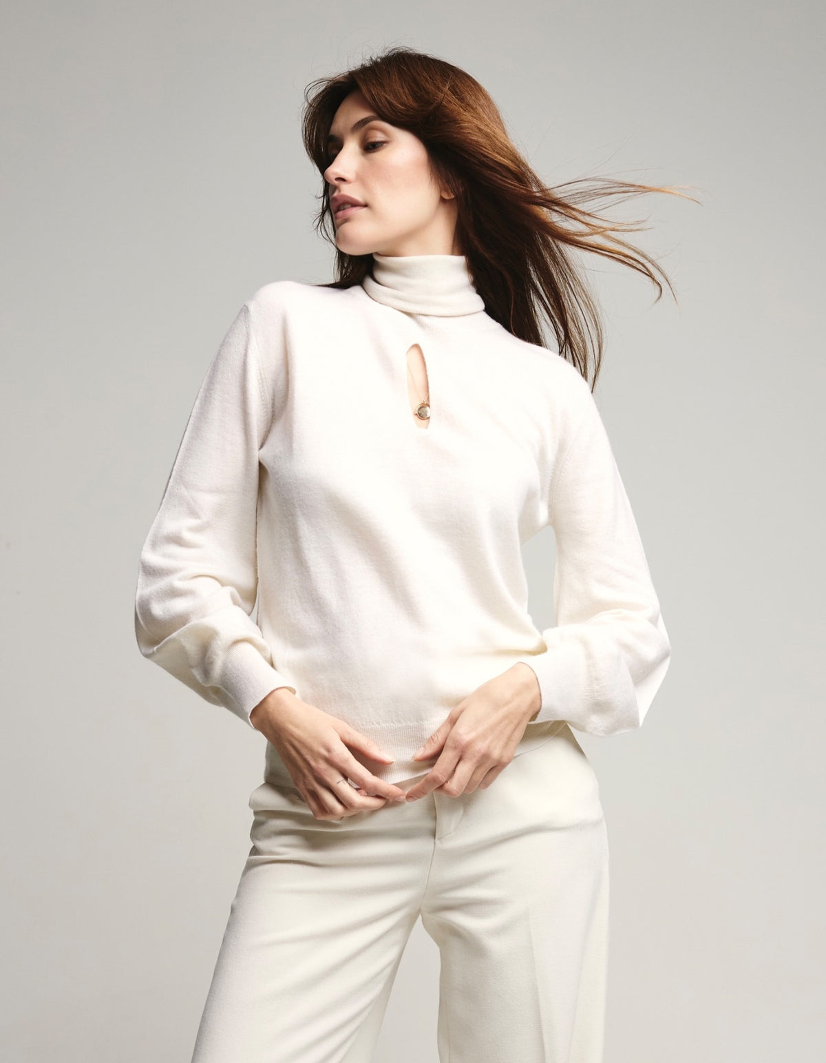 A woman modeling a cream-colored knit mock-neck sweater with a small keyhole cutout at the collar, paired with matching cream trousers, against a light gray studio background.