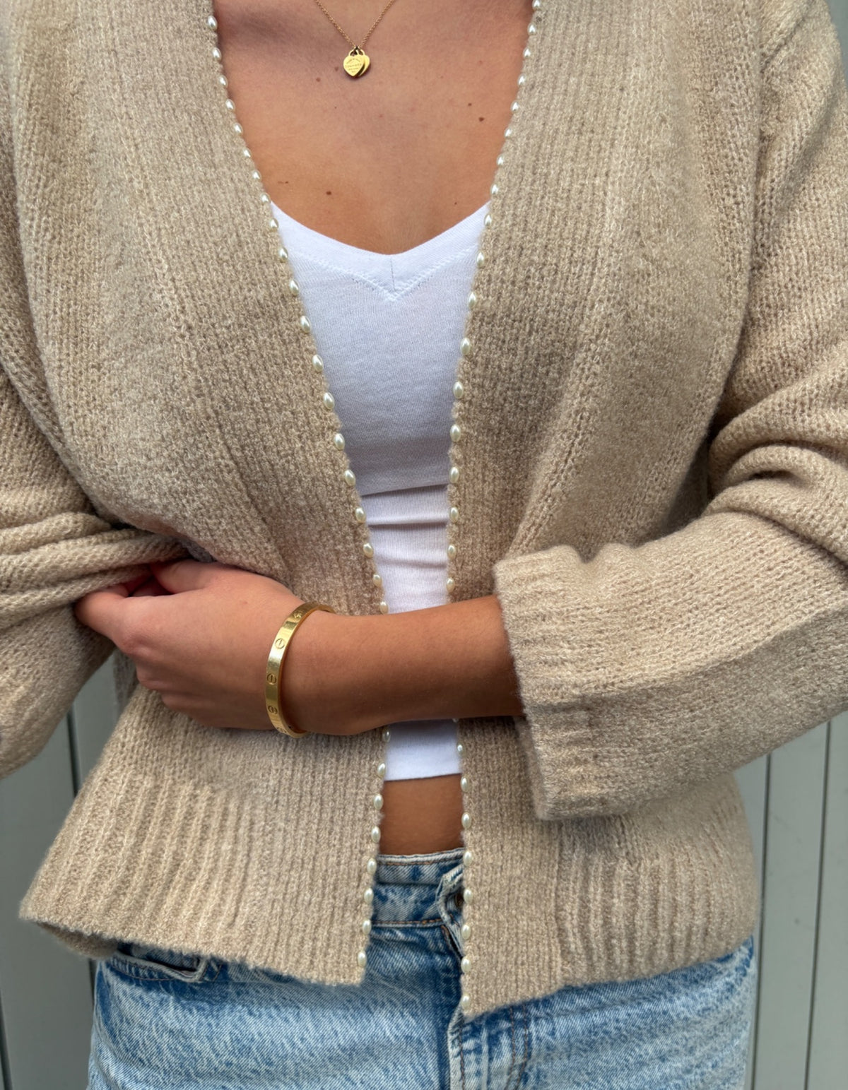 A person wearing a beige open-front knit cardigan with pearl-like trim over a white top and light blue jeans, shown from the shoulders to the hips.