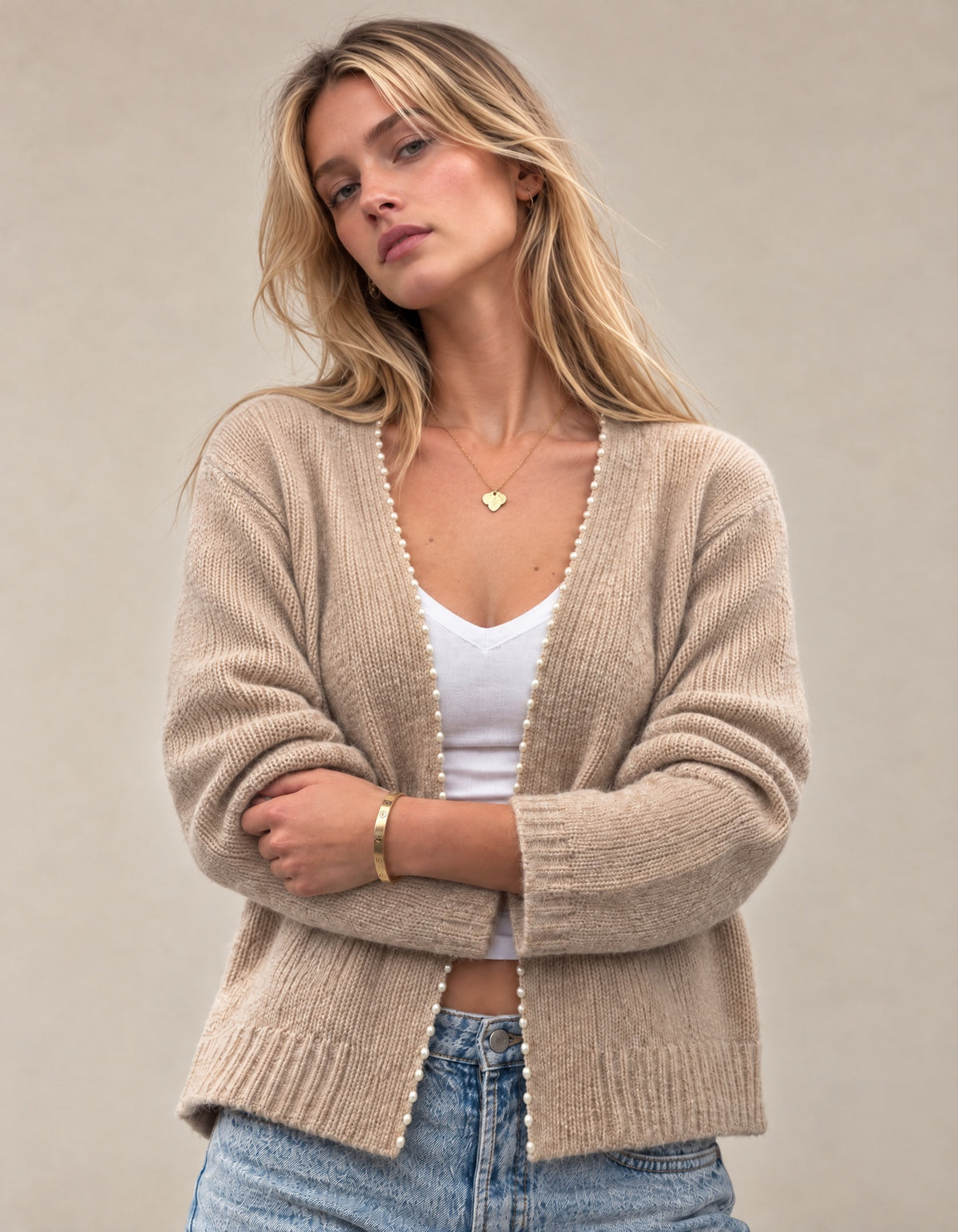 A woman wearing a beige knit cardigan with white pom-pom trim over a white tank top and blue jeans, standing against a neutral beige background.