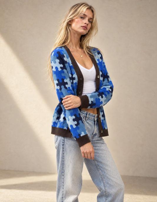 A woman models a blue and navy puzzle-piece pattern cardigan with dark brown ribbed trim, worn over a white tank top and light-wash jeans against a neutral beige background.