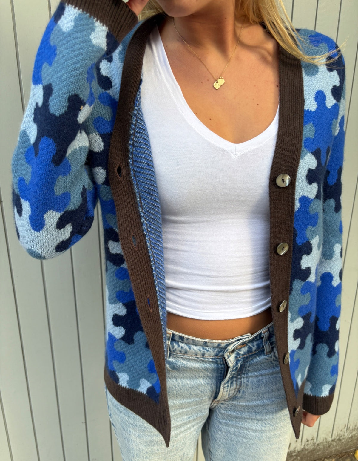 A person wearing a blue and navy puzzle-piece pattern knit cardigan with brown trim over a white crop top and light blue jeans, standing against a white paneled wall.