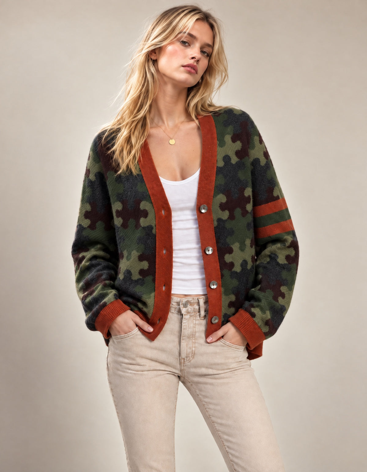 A woman models a green puzzle-piece camouflage cardigan with rust-brown trim and buttons, worn over a white tank top and light beige jeans against a neutral beige background.