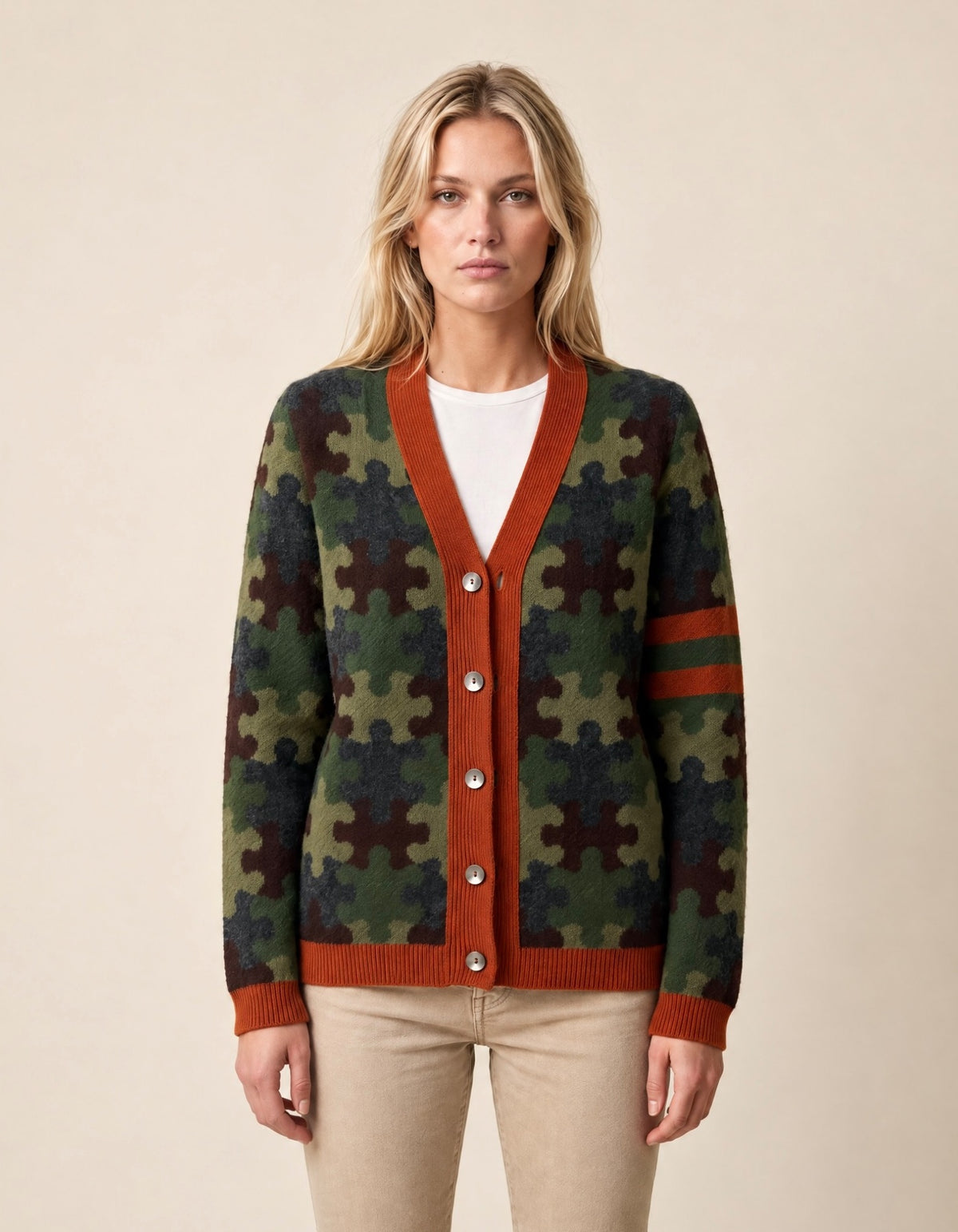 A woman modeling a cardigan sweater with a green and brown puzzle-piece pattern, rust-orange ribbed trim, and a white crewneck shirt underneath, paired with beige pants against a solid beige background.