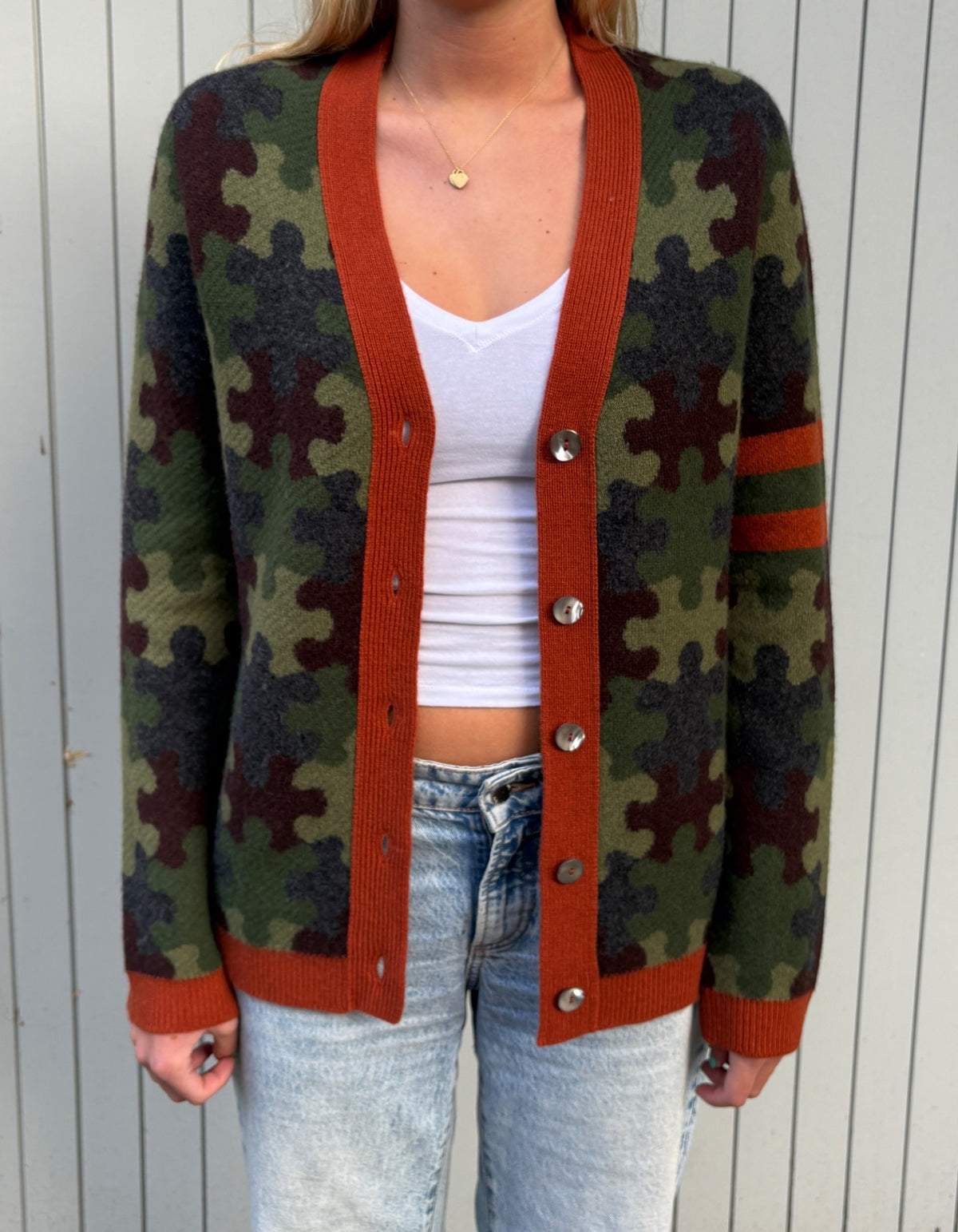 A person wearing a green and brown puzzle-piece camouflage cardigan with rust-orange ribbed trim over a white crop top and light-wash jeans, standing against a white paneled wall.