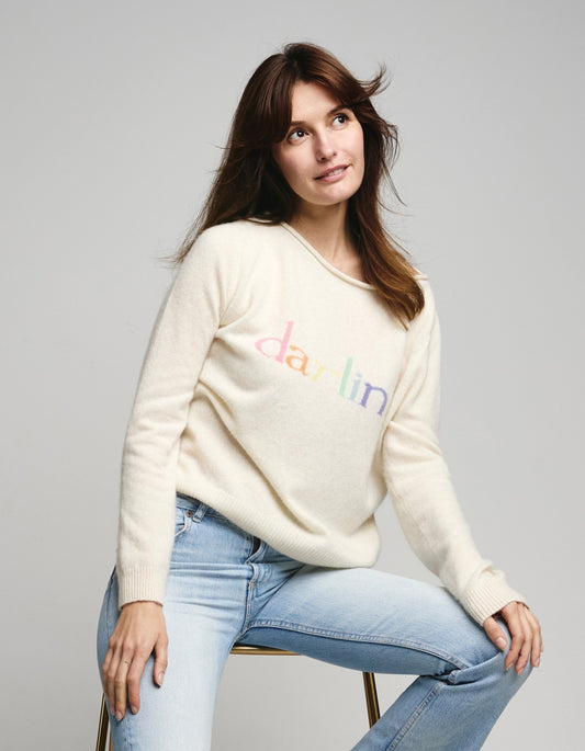 Woman wearing a cream sweater with colorful text darlin  and light blue jeans on a gray background